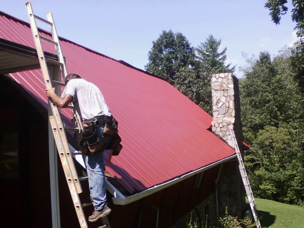 Roofer on a ladder beside a red metal roof and stone chimney — Soffit & Fascia Repair by Roofing Brothers in Lignum